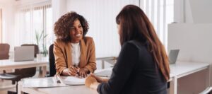 Two women sit at a desk in an office, smiling and having a discussion with documents in front of them.