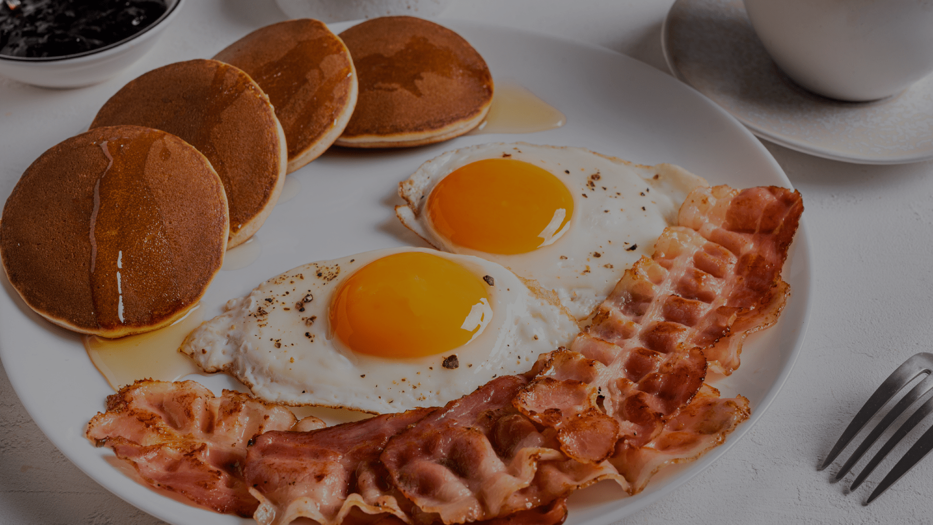 A breakfast plate with pancakes, fried eggs, and crispy bacon strips on a white table.