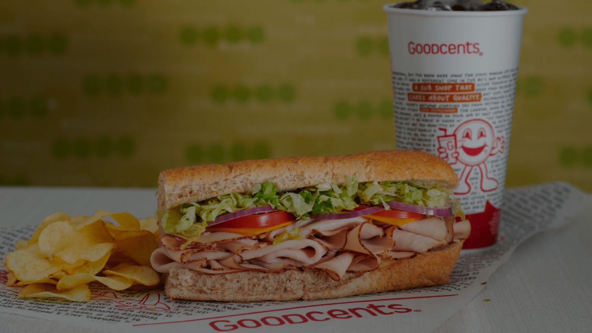 Sub sandwich with turkey, lettuce, tomato, and chips, next to a Goodcents drink cup on a table.