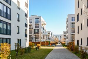 Modern apartment buildings under a clear blue sky with a central pathway surrounded by trimmed hedges and trees.