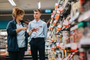 A supermarket manager training a young worker while standing next to shelves filled with products.