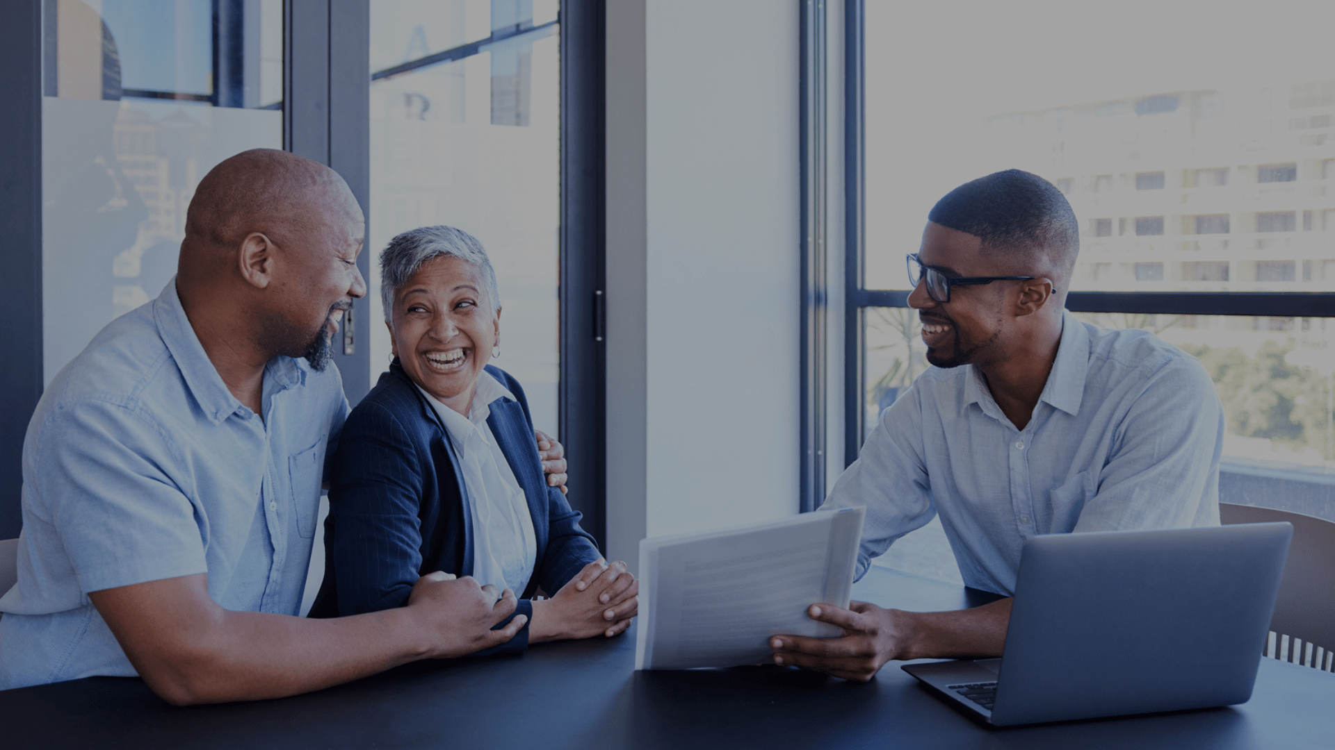 Three people sit at a table, smiling and talking, with documents and a laptop in front of them.