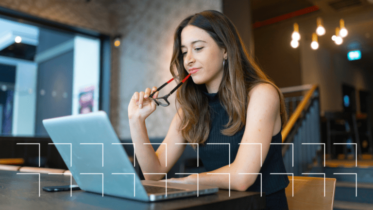 Woman sitting at a table, holding glasses, looking thoughtfully at a laptop in a modern workspace.
