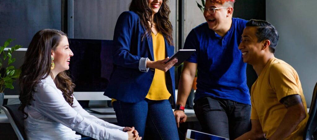 Four people smiling and talking in an office setting, with one person holding a tablet.