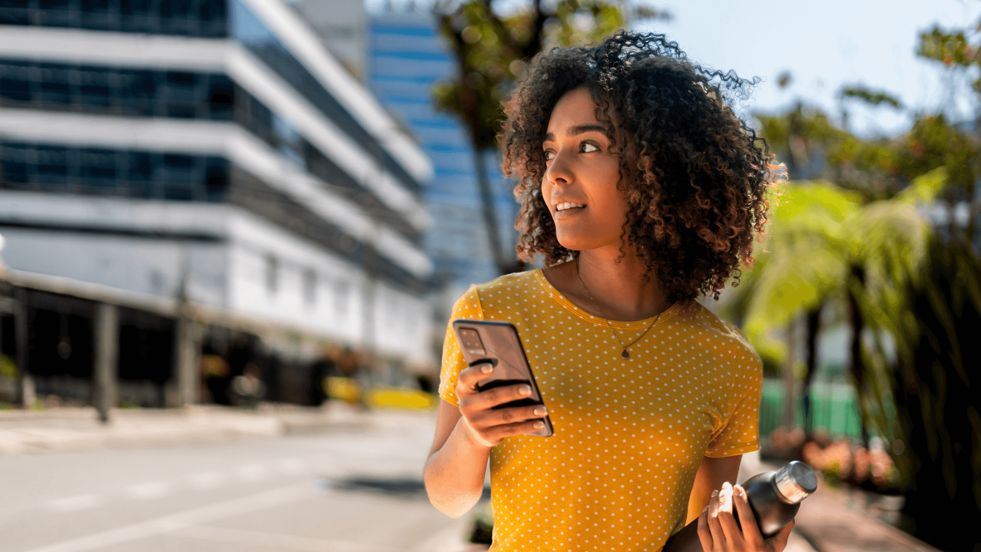 Woman in a yellow polka dot shirt holds a phone and water bottle, standing on a city street.