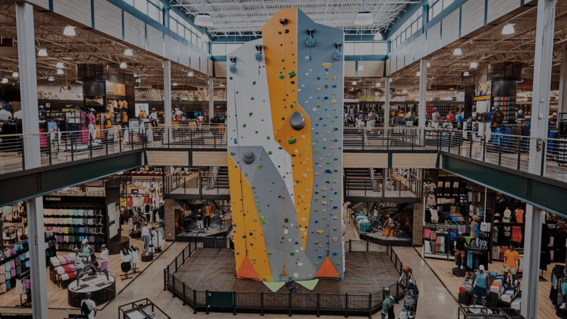 A large indoor climbing wall stands at the center of a sporting goods store with shoppers nearby.
