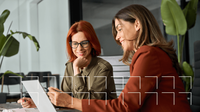 Two women smiling and working together at a desk with a laptop and documents in a modern office.