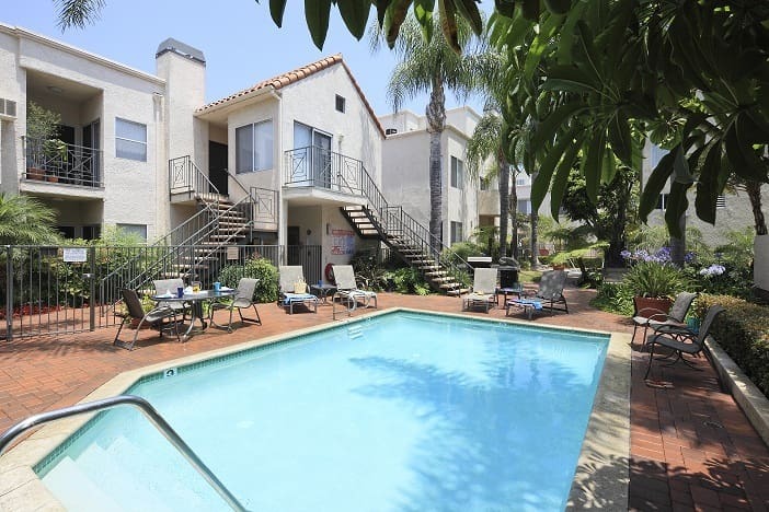 Outdoor swimming pool surrounded by lounge chairs and greenery in a modern apartment complex courtyard.