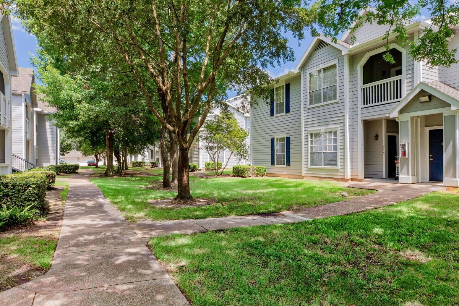 Sidewalks winding through grassy lawns and trees between white two-story apartment buildings on a sunny day.
