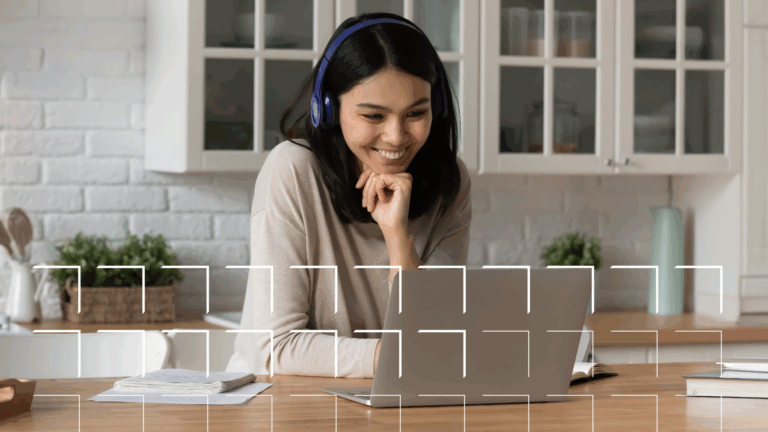 Woman wearing headphones, smiling while looking at a laptop in a bright kitchen with books on the table.