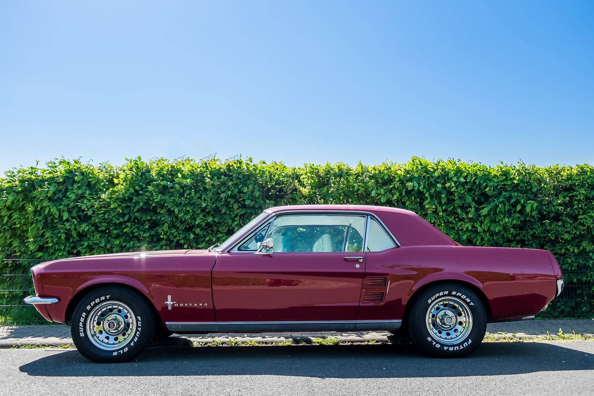 Side view of a classic red Ford Mustang parked on a street with green bushes and a clear blue sky behind.