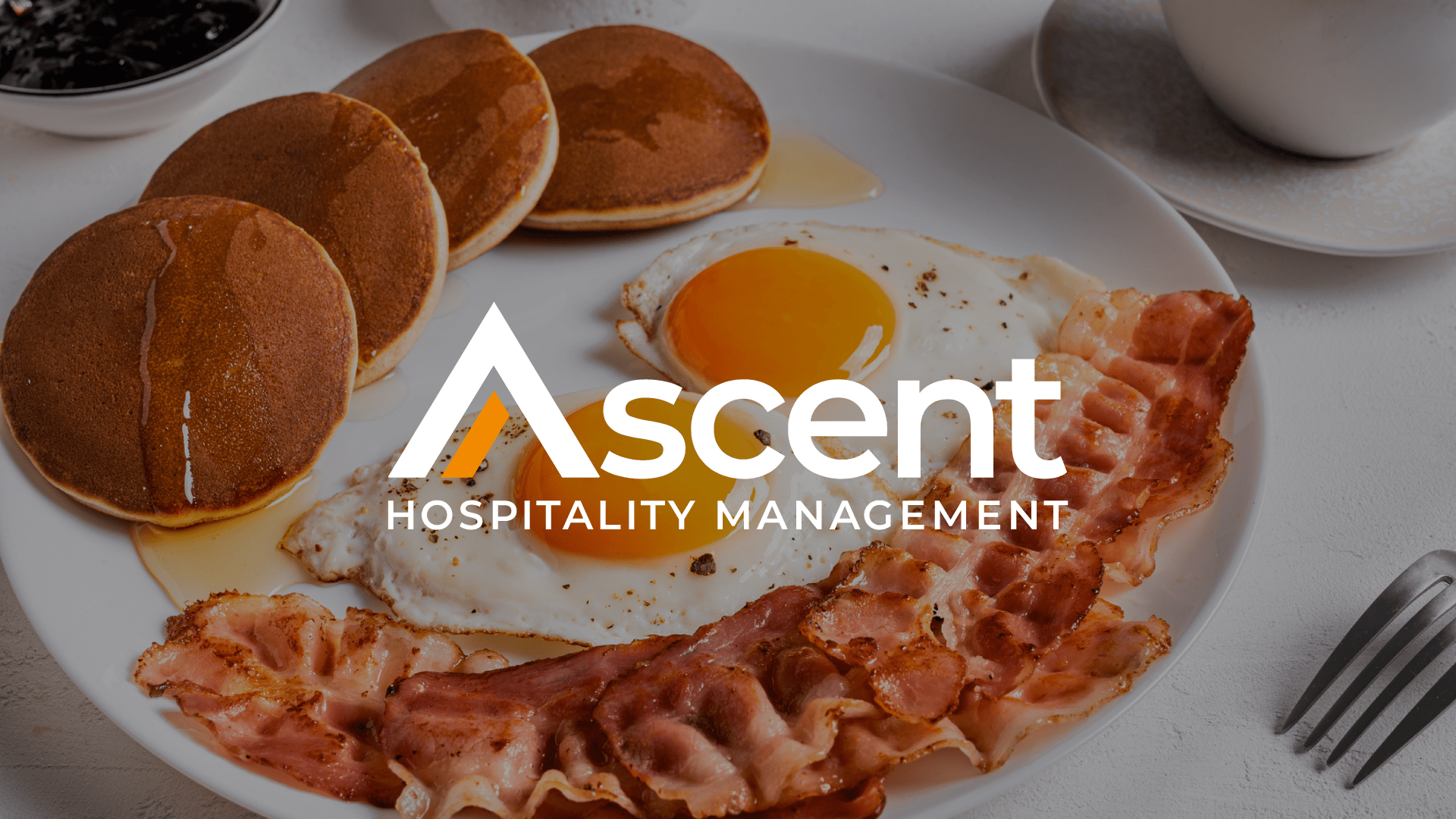 A breakfast plate with pancakes, a sunny-side-up egg, and bacon, overlaid with Ascent Hospitality Management.