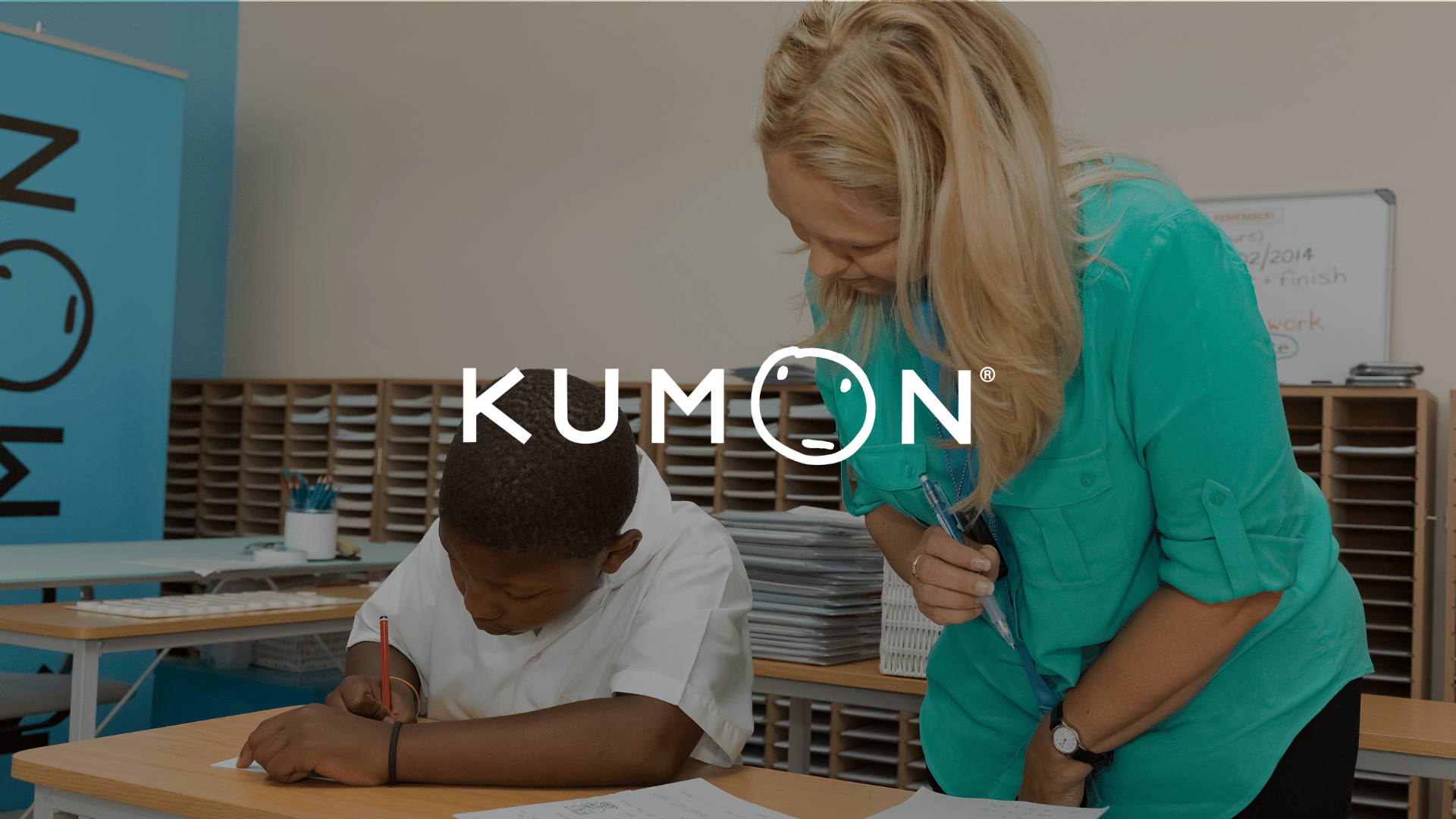 A teacher assists a student working at a desk in a classroom with the Kumon logo displayed over the image.