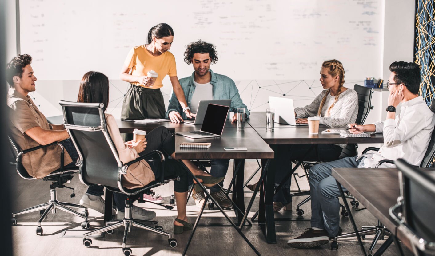 Six people have a meeting around a modern conference table with laptops, notebooks, and coffee cups.