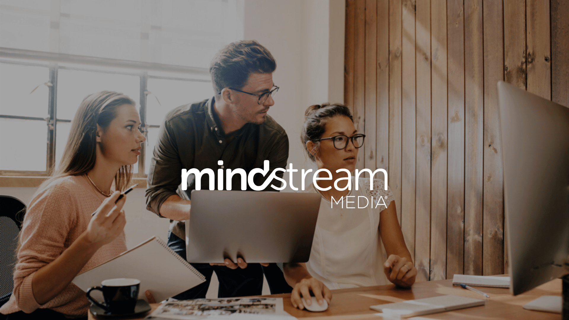 Three people collaborate at a desk with a computer; mindstream MEDIA logo is overlaid on the image.