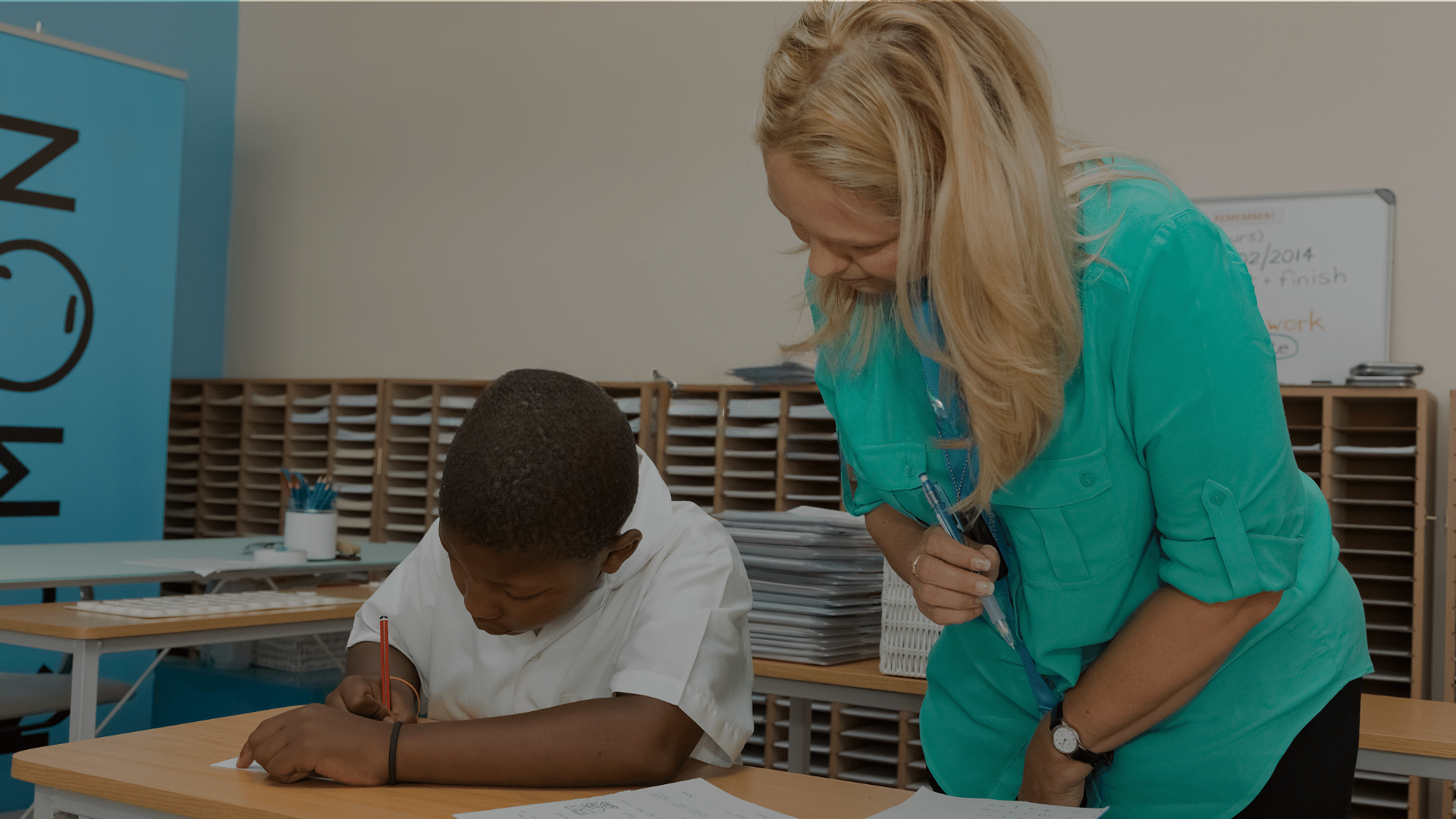 A teacher helps a student who is writing at a desk in a classroom.