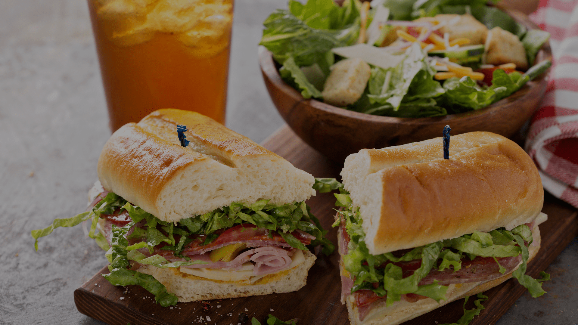 A sub sandwich with lettuce and meat, a bowl of salad, and a glass of iced tea on a table.